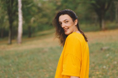 Parlak sarı bluzlu bir kadın, sonbaharda güneşin sıcaklığından ve doğanın güzelliğinden zevk alırken mutlu bir şekilde parkta yürür..