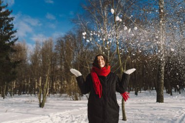 Kırmızı eşarplı gülümseyen bir kadın, parlak mavi gökyüzünün altındaki sakin bir ormanda karlı bir kış gününün tadını çıkarıyor. Noel ve Yeni Yıl Kutlama Konsepti.