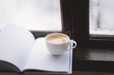 Sıcak bir fincan kahve pencerenin kenarında açık bir kitabın yanında durur, yanan bir mum yavaşça titreşir, karlı zemine karşı sakin bir atmosfer yaratır..