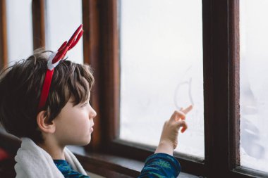 A young child wearing reindeer antlers sits wrapped in a cozy blanket, looking out a snow-covered window, evoking holiday spirit and warmth on a chilly day. clipart