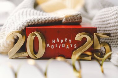 Mutlu yıllar 2025. Mutlu Tatiller mesajını gösteren canlı kırmızı bir hediye kutusu 2025 yılını gösteren altın rakamların altında duruyor..