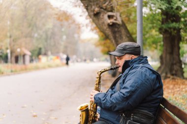 Bir saksafoncu, sonbaharda yapraklarla çevrili bir bankta otururken sakin bir parkta gösteri yapar.