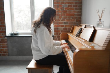 Kıvırcık saçlı bir kadın piyanonun başında oturmuş, güzel ışıklandırılmış oturma odasında yarattığı müziğin keyfini çıkarıyor..