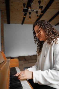 Kıvırcık saçlı bir kadın piyanonun başında oturmuş, güzel ışıklandırılmış oturma odasında yarattığı müziğin keyfini çıkarıyor..