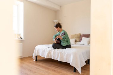 Sıcak bir odada gitar çalan bir kadın. Güneş ışığı yumuşak gölgeler bırakır, müzikal ifadesi için sakin bir atmosfer yaratır..