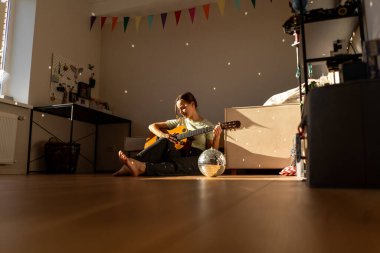 Sıcak bir odada gitar çalan bir kadın. Güneş ışığı yumuşak gölgeler bırakır, müzikal ifadesi için sakin bir atmosfer yaratır..