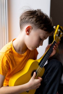 Turuncu gömlekli bir çocuk evdeki odasında parlak sarı bir ukulele tıngırdatmaya odaklanmış..