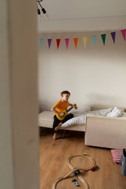 Turuncu gömlekli bir çocuk evdeki odasında parlak sarı bir ukulele tıngırdatmaya odaklanmış..