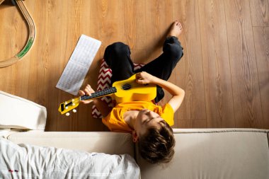 Turuncu gömlekli bir çocuk evdeki odasında parlak sarı bir ukulele tıngırdatmaya odaklanmış..