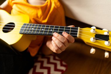 Turuncu gömlekli bir çocuk evdeki odasında parlak sarı bir ukulele tıngırdatmaya odaklanmış..