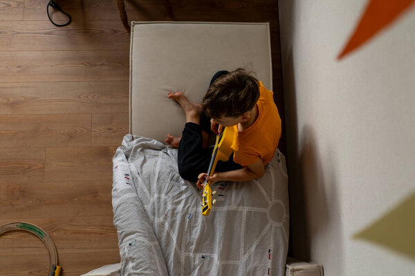 A boy dressed in an orange shirt focused on strumming a bright yellow ukulele in a cozy his room at home.
