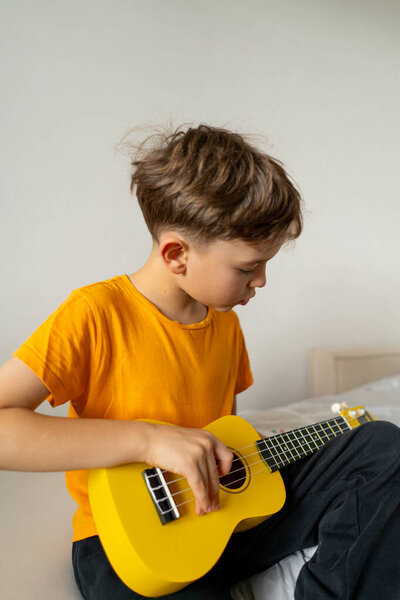 A boy dressed in an orange shirt focused on strumming a bright yellow ukulele in a cozy his room at home.