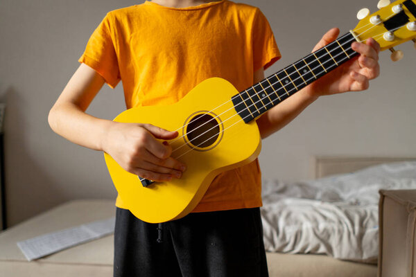 A boy dressed in an orange shirt focused on strumming a bright yellow ukulele in a cozy his room at home.