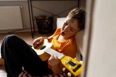 Turuncu gömlekli bir çocuk evdeki odasında parlak sarı bir ukulele tıngırdatmaya odaklanmış..