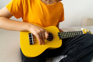 Turuncu gömlekli bir çocuk evdeki odasında parlak sarı bir ukulele tıngırdatmaya odaklanmış..