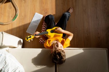 Turuncu gömlekli bir çocuk evdeki odasında parlak sarı bir ukulele tıngırdatmaya odaklanmış..