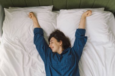 Bir kadın yatakta sırt üstü uzanır, mavi bir gömlek giyer, esnerken gülümser. Güneş ışığı onun huzurlu ifadesini nazikçe aydınlatıyor, sakin bir sabah atmosferini güçlendiriyor..