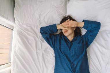 Bir kadın yatakta sırt üstü uzanır, mavi bir gömlek giyer, esnerken gülümser. Güneş ışığı onun huzurlu ifadesini nazikçe aydınlatıyor, sakin bir sabah atmosferini güçlendiriyor..