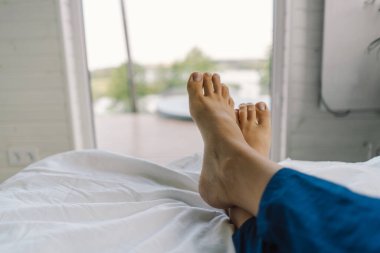 Çıplak ayaklar temiz beyaz bir yatak örtüsünde görülebilir, bu da rahat bir sabahı gösterir. Yumuşak ışık uzayın sükunetini vurgular, barışçıl bir atmosfer yaratır..