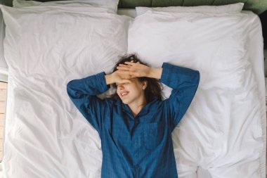 Bir kadın yatakta sırt üstü uzanır, mavi bir gömlek giyer, esnerken gülümser. Güneş ışığı onun huzurlu ifadesini nazikçe aydınlatıyor, sakin bir sabah atmosferini güçlendiriyor..