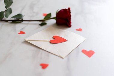 Masanın üzerinde kalp şeklinde konfeti ve kırmızı bir gülle süslenmiş beyaz bir zarf var. Özel bir günde aşkı ifade etmek için mükemmel bir ortam. Sevgililer Günün kutlu olsun.