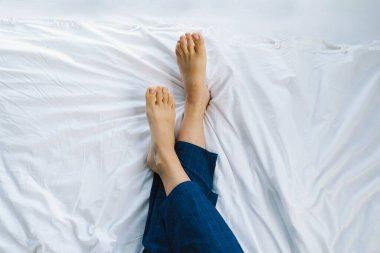 Çıplak ayaklar temiz beyaz bir yatak örtüsünde görülebilir, bu da rahat bir sabahı gösterir. Yumuşak ışık uzayın sükunetini vurgular, barışçıl bir atmosfer yaratır..