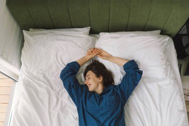 Bir kadın yatakta sırt üstü uzanır, mavi bir gömlek giyer, esnerken gülümser. Güneş ışığı onun huzurlu ifadesini nazikçe aydınlatıyor, sakin bir sabah atmosferini güçlendiriyor..