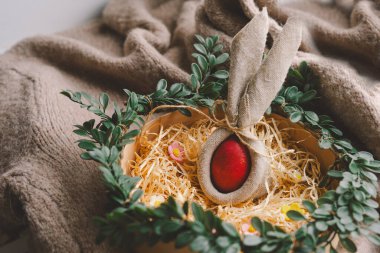 Paskalya için dekoratif bir yuva, yumuşak samanların içine yerleştirilmiş ve dokulu bir battaniyenin üzerindeki yeşil yapraklarla çevrili çiçek aksanlı renkli yumurtalar sergiler. Paskalya konsepti