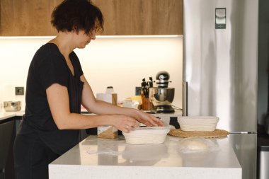 Eller, temiz mutfak tezgahına un serpiştirilmiş hamuru ustalıkla sarar ve şekillendirir. Sağlıklı mayalı ekmek serpiştirir. Parlak alan sıcak bir fırın atmosferini işaret ediyor..