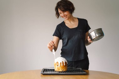 Bir kadın Paskalya panettonunu evde yapılacak bahar kutlamasına hazırlanırken beyaz krema ve renkli çikolata parçacıklarıyla süslüyor. Taze çiçekler ayara güzellik katar..