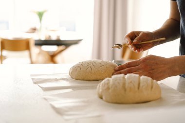 Woman hands skillfully score the surface of a round dough loaf, preparing sourdough bread for baking. The bright kitchen setting provides a warm atmosphere and fresh ingredients. clipart