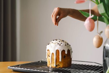 Bir kadın Paskalya panettonunu evde yapılacak bahar kutlamasına hazırlanırken beyaz krema ve renkli çikolata parçacıklarıyla süslüyor. Taze çiçekler ayara güzellik katar..