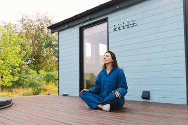 A woman is sitting cross-legged on a wooden deck, practicing meditation. The serene atmosphere is enhanced by the lush greenery around a contemporary blue residence. clipart