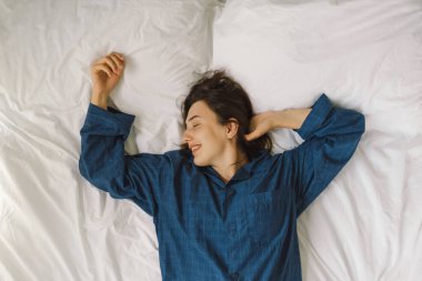 Bir kadın yatakta sırt üstü uzanır, mavi bir gömlek giyer, esnerken gülümser. Güneş ışığı onun huzurlu ifadesini nazikçe aydınlatıyor, sakin bir sabah atmosferini güçlendiriyor..