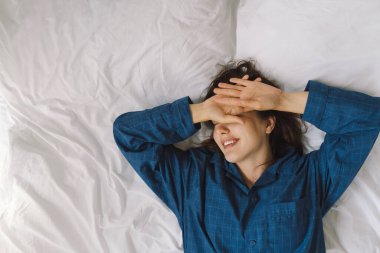 Bir kadın yatakta sırt üstü uzanır, mavi bir gömlek giyer, esnerken gülümser. Güneş ışığı onun huzurlu ifadesini nazikçe aydınlatıyor, sakin bir sabah atmosferini güçlendiriyor..