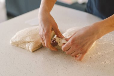 Eller, temiz mutfak tezgahına un serpiştirilmiş hamuru ustalıkla sarar ve şekillendirir. Sağlıklı mayalı ekmek serpiştirir. Parlak alan sıcak bir fırın atmosferini işaret ediyor..