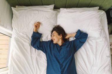 Bir kadın yatakta sırt üstü uzanır, mavi bir gömlek giyer, esnerken gülümser. Güneş ışığı onun huzurlu ifadesini nazikçe aydınlatıyor, sakin bir sabah atmosferini güçlendiriyor..