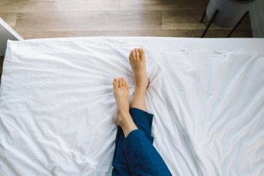 Çıplak ayaklar temiz beyaz bir yatak örtüsünde görülebilir, bu da rahat bir sabahı gösterir. Yumuşak ışık uzayın sükunetini vurgular, barışçıl bir atmosfer yaratır..