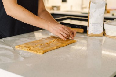 Günlük giysiler içindeki bir kadın hamur işini hazırlar. Memnun ve odaklanmış görünüyor, ışık dolu bir mutfakta pişirme sürecinin tadını çıkarıyor..