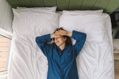 Bir kadın yatakta sırt üstü uzanır, mavi bir gömlek giyer, esnerken gülümser. Güneş ışığı onun huzurlu ifadesini nazikçe aydınlatıyor, sakin bir sabah atmosferini güçlendiriyor..