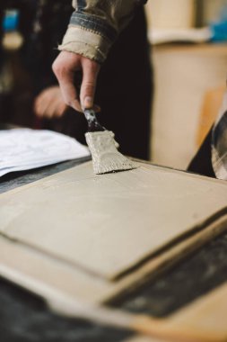 Elinde fırça olan bir kız kille çalışır. Çalışma alanı, doğal ışık tarafından aydınlatılan tamamlanmamış parça ve araçlarla dolu..