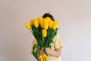 Bir çocuk elinde büyük bir buket sarı lale ile evde duruyor, yüzünde kocaman bir gülümsemeyle mutluluk saçıyor. Parlak atmosfer neşe ve sıcaklığı yansıtır. Anneler Günü ve Bahar Konsepti