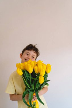 Bir çocuk elinde büyük bir buket sarı lale ile evde duruyor, yüzünde kocaman bir gülümsemeyle mutluluk saçıyor. Parlak atmosfer neşe ve sıcaklığı yansıtır. Anneler Günü ve Bahar Konsepti