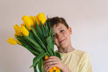 Bir çocuk elinde büyük bir buket sarı lale ile evde duruyor, yüzünde kocaman bir gülümsemeyle mutluluk saçıyor. Parlak atmosfer neşe ve sıcaklığı yansıtır. Anneler Günü ve Bahar Konsepti