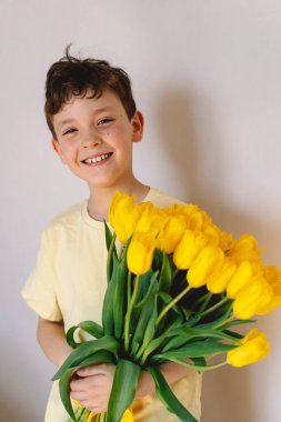Bir çocuk elinde büyük bir buket sarı lale ile evde duruyor, yüzünde kocaman bir gülümsemeyle mutluluk saçıyor. Parlak atmosfer neşe ve sıcaklığı yansıtır. Anneler Günü ve Bahar Konsepti
