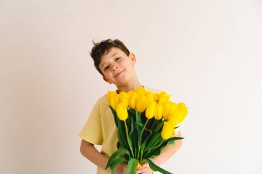 Bir çocuk elinde büyük bir buket sarı lale ile evde duruyor, yüzünde kocaman bir gülümsemeyle mutluluk saçıyor. Parlak atmosfer neşe ve sıcaklığı yansıtır. Anneler Günü ve Bahar Konsepti