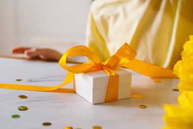 Bir çocuk mutfak masasında oturmuş, anneler günü hediyesinde fiyonk bağlıyor. Güzel sarı laleler onu sarıp, neşeli bir atmosfer yaratıyor..