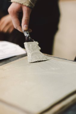 Elinde fırça olan bir kız kille çalışır. Çalışma alanı, doğal ışık tarafından aydınlatılan tamamlanmamış parça ve araçlarla dolu..