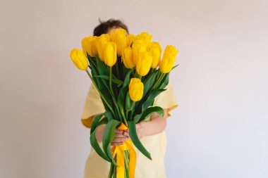 Bir çocuk elinde büyük bir buket sarı lale ile evde duruyor, yüzünde kocaman bir gülümsemeyle mutluluk saçıyor. Parlak atmosfer neşe ve sıcaklığı yansıtır. Anneler Günü ve Bahar Konsepti