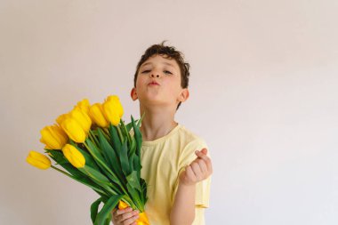 Bir çocuk elinde büyük bir buket sarı lale ile evde duruyor, yüzünde kocaman bir gülümsemeyle mutluluk saçıyor. Parlak atmosfer neşe ve sıcaklığı yansıtır. Anneler Günü ve Bahar Konsepti